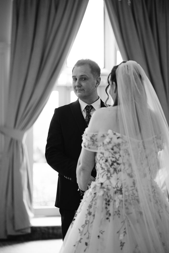 A groom in a suit stands facing a bride in an off-shoulder floral wedding dress and veil, indoors near large curtained windows, capturing the romantic ambience of a Late Summer Wedding.