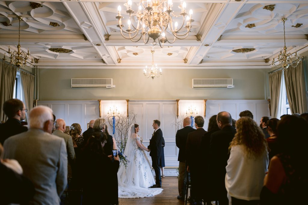 A bride and groom stand facing each other at the altar in a formal late summer wedding ceremony, surrounded by guests in a beautifully decorated room at The Maynard, with chandeliers and ornate ceiling details.