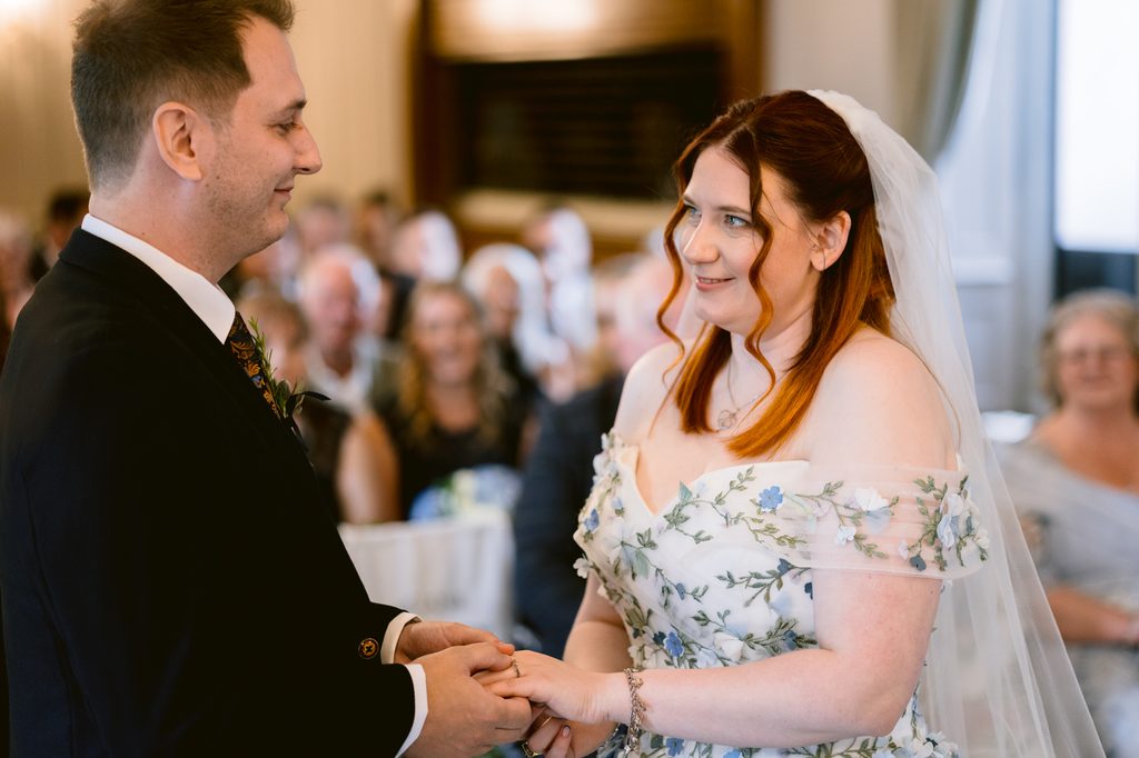 Bride and groom stand facing each other, holding hands during their late summer wedding ceremony at The Maynard, with guests seated and watching in the blurred Padley Gorge background.