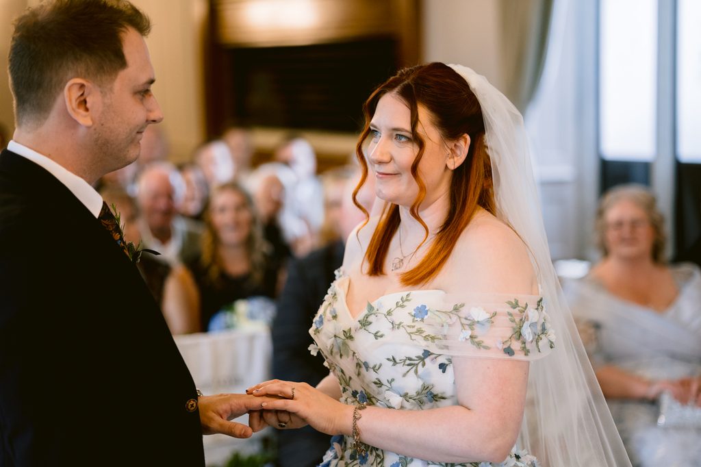 A bride in a floral dress and veil places a ring on the groom’s finger during a late summer wedding ceremony at The Maynard, with guests seated in the background and the beauty of Padley Gorge nearby.