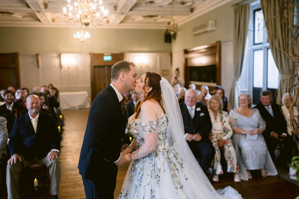 A bride and groom kiss at the altar during their wedding ceremony at The Maynard, in a room filled with seated guests, capturing the romance of a late summer wedding.