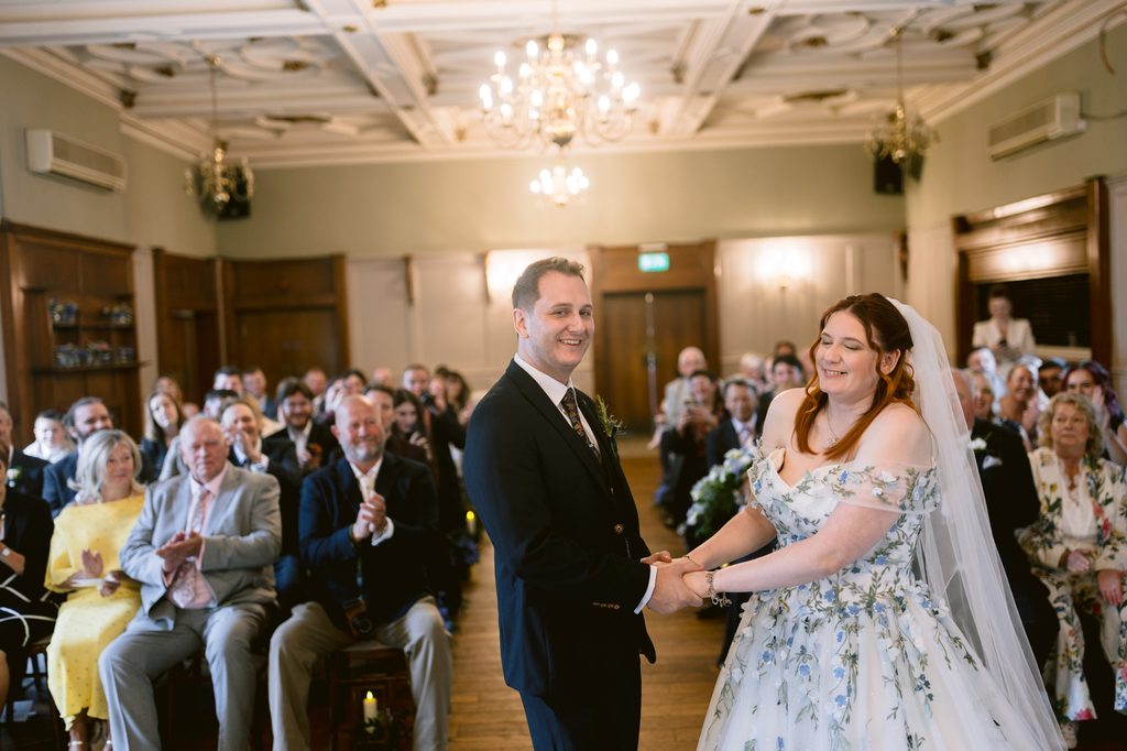 A bride and groom hold hands and smile at their romantic indoor wedding ceremony at The Maynard, with guests seated and watching in the background, capturing the magic of their late summer wedding.