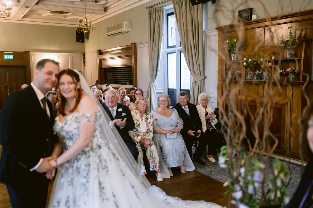 A bride and groom stand at the altar while guests seated behind them watch the wedding ceremony in a beautifully decorated indoor venue at The Maynard, capturing the charm of a late summer wedding.