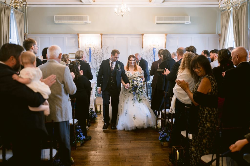 A bride and groom walk down the aisle together in a beautifully decorated indoor venue at The Maynard during their late summer wedding, whilst guests on both sides stand and applaud.