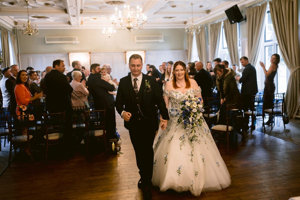 A bride and groom walk down the aisle holding hands in a beautifully decorated indoor venue at The Maynard, celebrating their late summer wedding as guests stand and watch in the background.