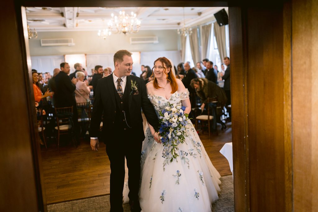 A bride and groom walk into The Maynard’s reception hall filled with seated guests. The bride wears a white floral gown and carries a bouquet; the groom wears a dark suit, perfectly capturing the elegance of their late summer wedding.