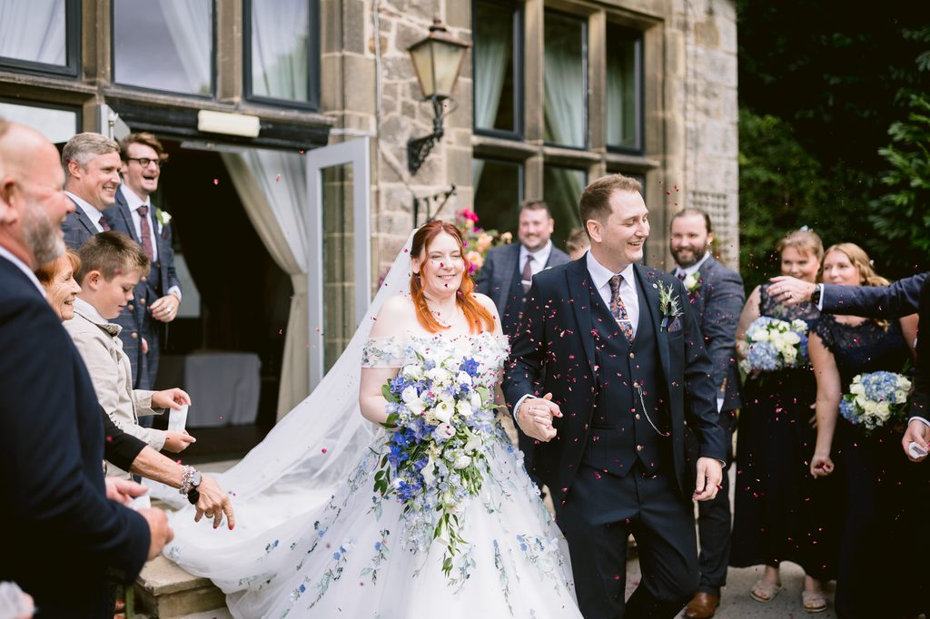 A bride and groom walk outside the stone building at The Maynard, smiling and holding hands, as guests throw confetti and celebrate around them during their late summer wedding.