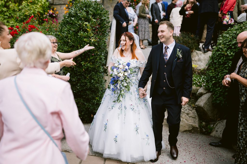 A bride and groom holding hands walk outdoors at Padley Gorge, smiling, surrounded by guests in formal attire. The bride carries a large bouquet with blue flowers, capturing the joyful spirit of their late summer wedding.