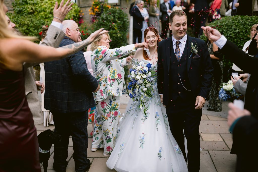 A bride and groom walk outdoors among guests throwing confetti, surrounded by greenery and flowers during their late summer wedding. The bride wears a white dress with blue accents and holds a matching bouquet, as the celebration unfolds at The Maynard near picturesque Padley Gorge.