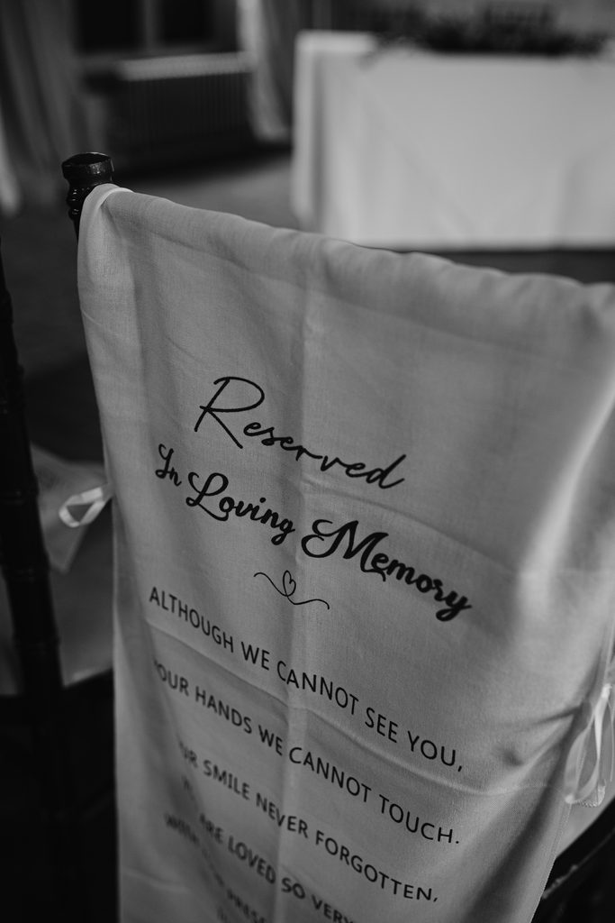 A white chair cover reads “Reserved In Loving Memory” with a poem, set on a dark chair in a softly lit room at The Maynard—capturing the gentle elegance of a late summer wedding.