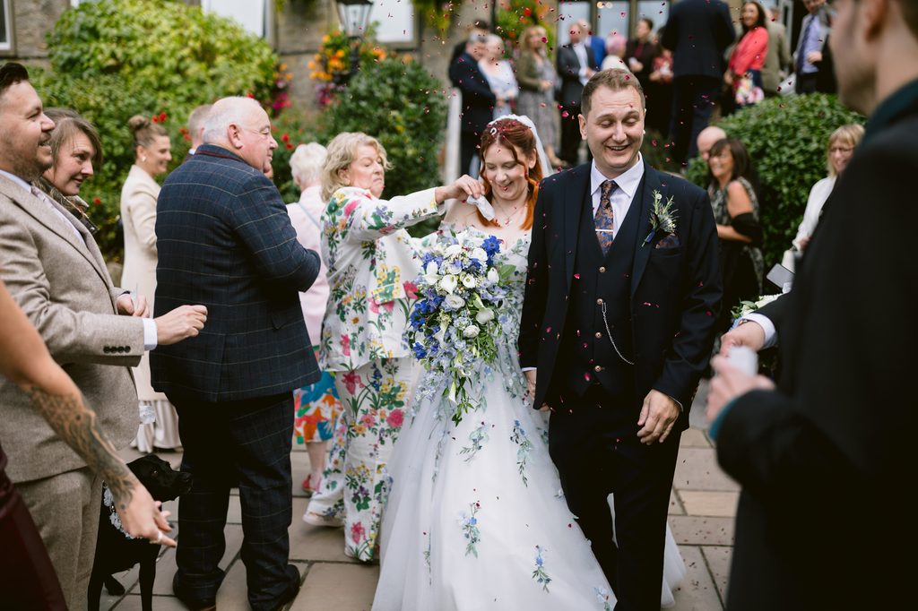 A bride and groom walk outside among guests throwing confetti during their late summer wedding at The Maynard, with the bride holding a bouquet of white and blue flowers and people smiling around them.