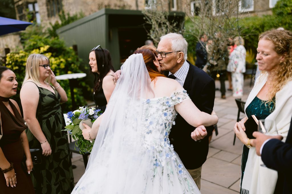 An older man kisses a bride in a white floral dress and veil while wedding guests stand and watch in an outdoor setting at The Maynard, capturing the warmth of a late summer wedding near Padley Gorge.
