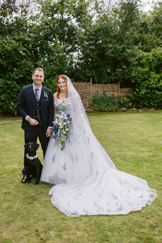 A bride in a white gown and groom in a dark suit stand on grass with a black dog wearing a bow tie, against a backdrop of trees and a wooden fence, capturing the magic of their late summer wedding near Padley Gorge at The Maynard.