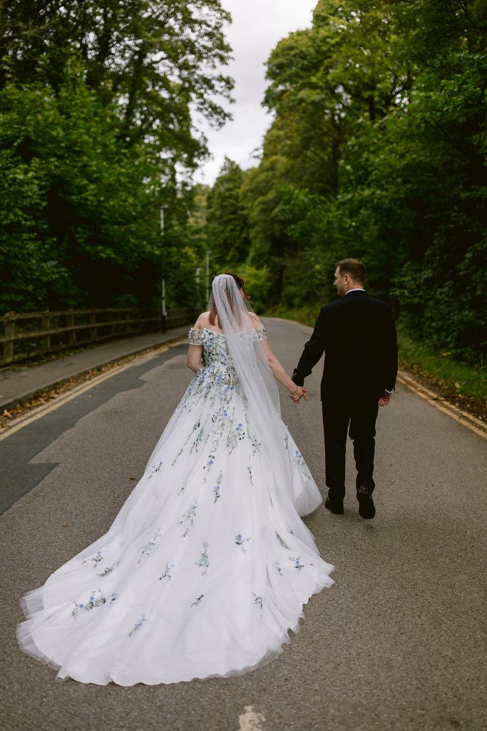 A bride and groom walk hand in hand down a tree-lined road, the bride wearing a long white gown with a veil, and the groom in a dark suit, capturing the magic of their late summer wedding near Padley Gorge.