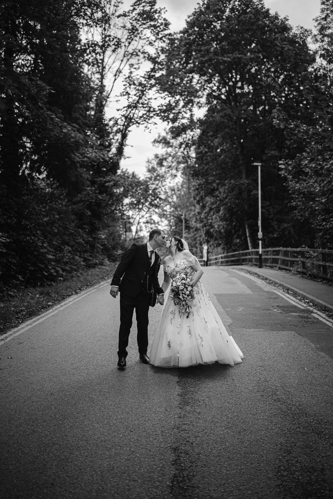 A bride and groom kiss whilst standing in the middle of a tree-lined road near Padley Gorge, dressed in formal wedding attire, capturing the romance of a late summer wedding at The Maynard.