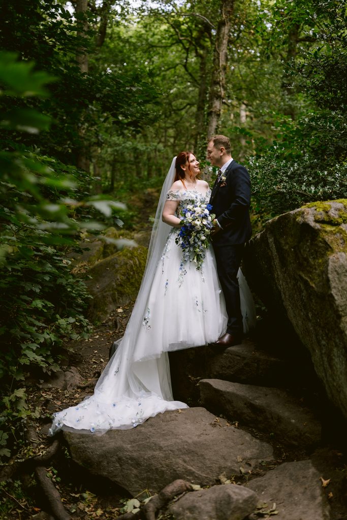 A bride and groom stand together outdoors on rocks in Padley Gorge, gazing at each other. The bride holds a blue and white bouquet and wears a long white dress with a veil, capturing the enchanting atmosphere of a late summer wedding.