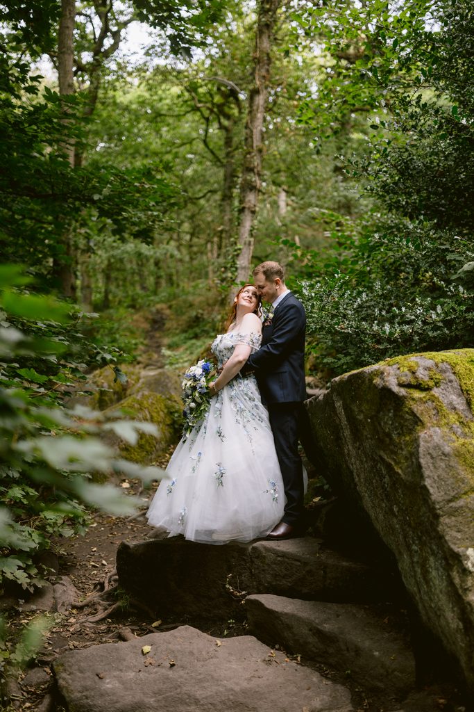 A bride and groom stand on rocks in the lush, green forest of Padley Gorge. The bride, dressed in a white gown with floral details and holding a bouquet, is radiant for their late summer wedding. The groom stands behind her in a dark suit, completing this beautiful scene before they celebrate at The Maynard.
