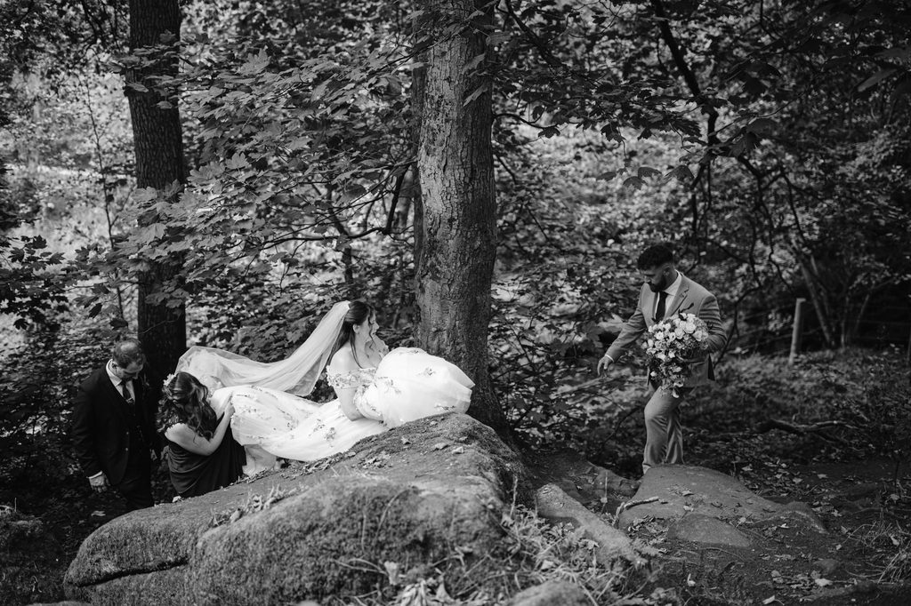 A bride in a wedding dress climbs rocks in the wooded beauty of Padley Gorge, assisted by two people, while another person in a suit follows carrying a bouquet—capturing the adventurous spirit of a late summer wedding near The Maynard.