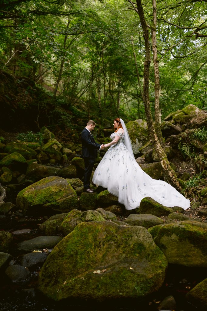 A bride and groom stand together on mossy rocks in a lush, green forest at Padley Gorge, holding hands and facing each other, capturing the enchanting essence of a late summer wedding.