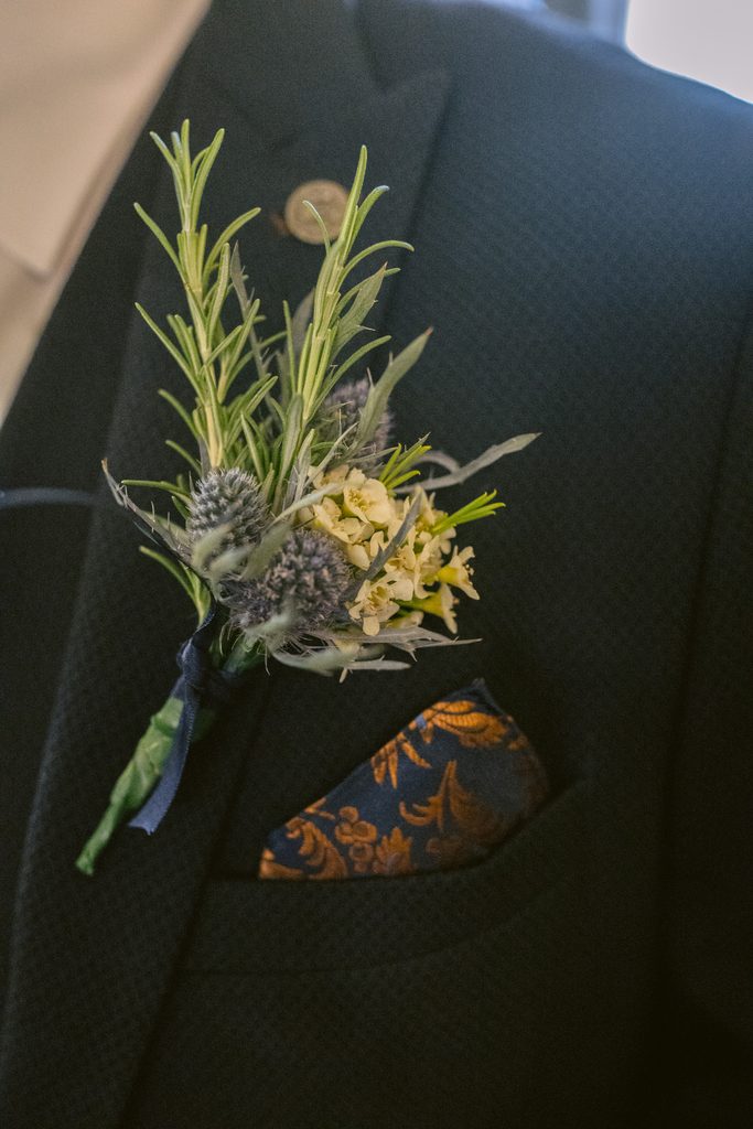 A close-up of a suit jacket with a gold-patterned pocket square and a buttonhole made of greenery and small yellow and purple flowers, perfect for a late summer wedding at The Maynard.