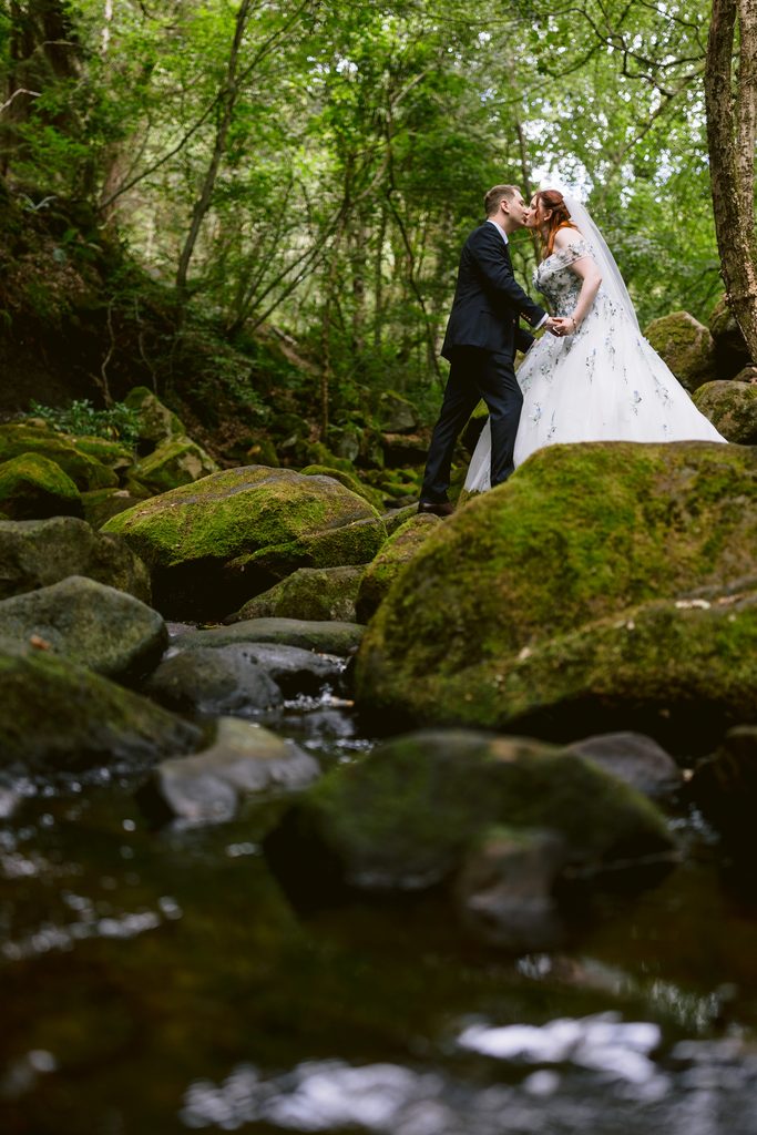 A bride and groom stand on mossy rocks by a stream in Padley Gorge, sharing an intimate moment surrounded by greenery. This enchanting late summer wedding scene captures the magic of The Maynard nestled within a tranquil, wooded area.