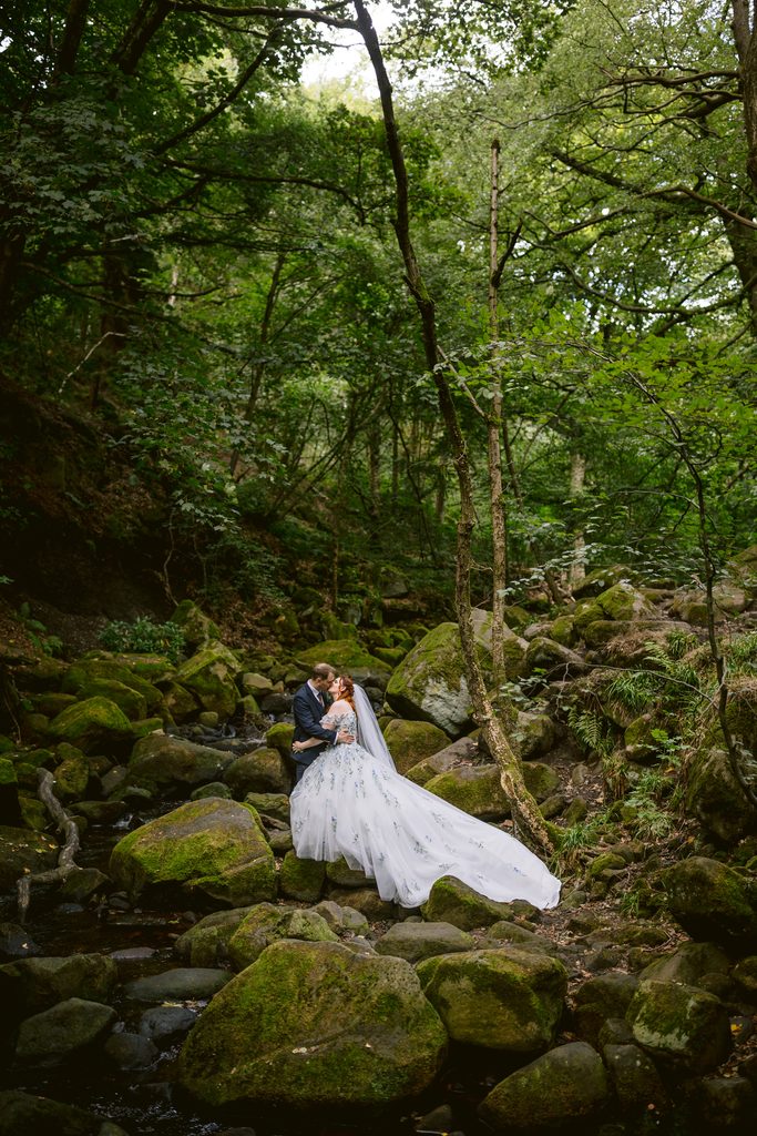 A bride and groom stand on mossy rocks in Padley Gorge, embracing. The bride wears a white dress with a long veil, and the groom is in a dark suit. Lush green trees surround them, capturing the enchanting spirit of a late summer wedding.