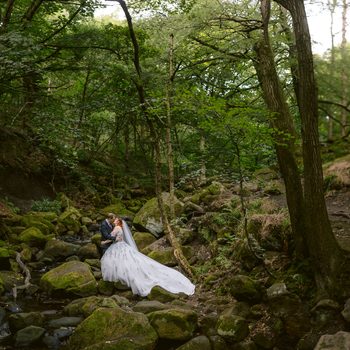 A bride and groom stand on mossy rocks in a forest at Padley Gorge, embracing near a small stream, surrounded by tall trees and greenery—a magical moment from their late summer wedding at The Maynard.