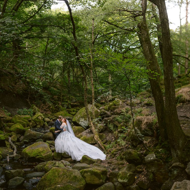 A bride and groom stand on mossy rocks in a forest at Padley Gorge, embracing near a small stream, surrounded by tall trees and greenery—a magical moment from their late summer wedding at The Maynard.