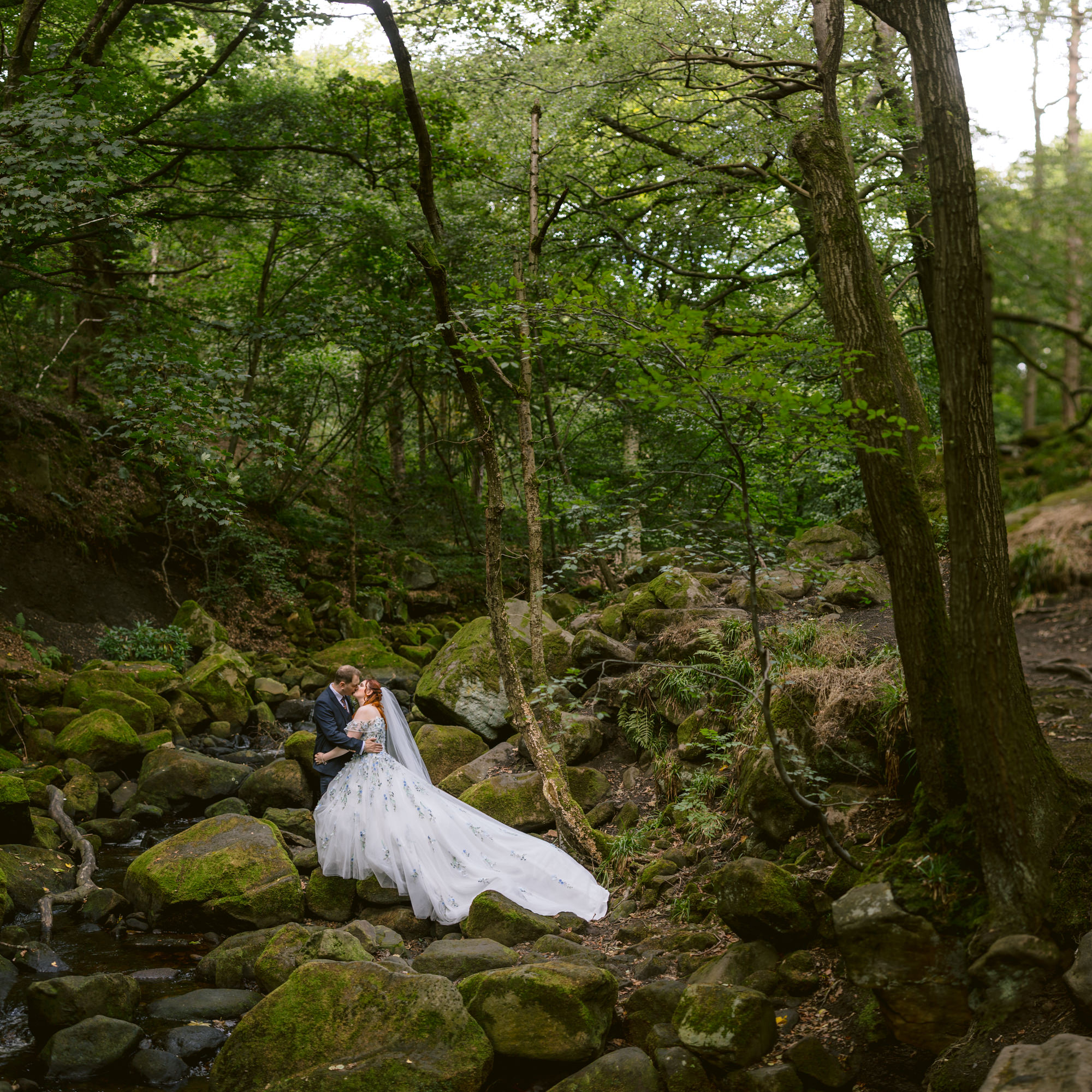 A bride and groom stand on mossy rocks in a forest at Padley Gorge, embracing near a small stream, surrounded by tall trees and greenery—a magical moment from their late summer wedding at The Maynard.