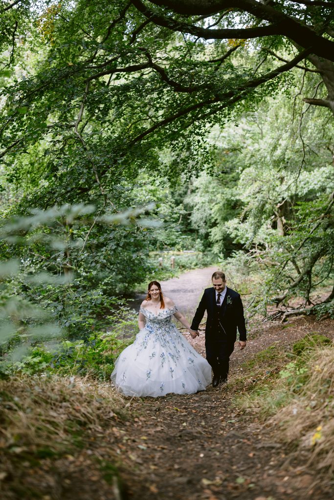 A bride in a white dress and a groom in a black suit walk hand in hand along a wooded path surrounded by trees, capturing the romance of their late summer wedding at Padley Gorge near The Maynard.