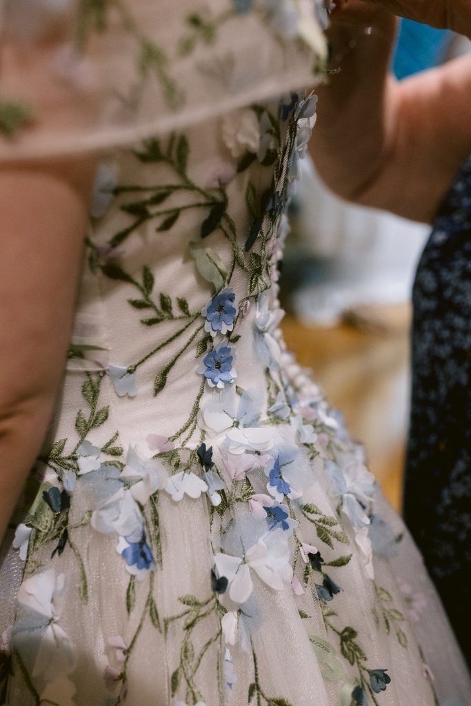 Close-up of a cream-coloured dress with blue and white floral appliqués and green embroidered leaves, worn by a person being assisted, perfect for a late summer wedding at The Maynard or an enchanting celebration near Padley Gorge.