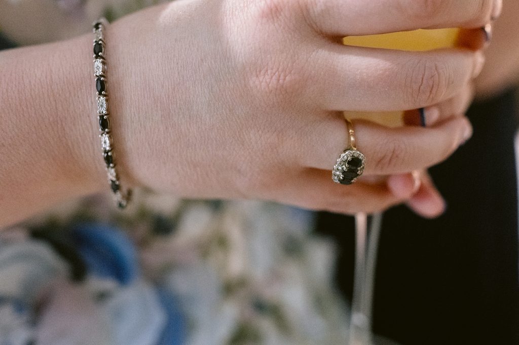 A hand wearing a black and silver bracelet and an ornate ring holds a glass with a yellow drink inside, capturing the relaxed elegance of a Late Summer Wedding at The Maynard near Padley Gorge.