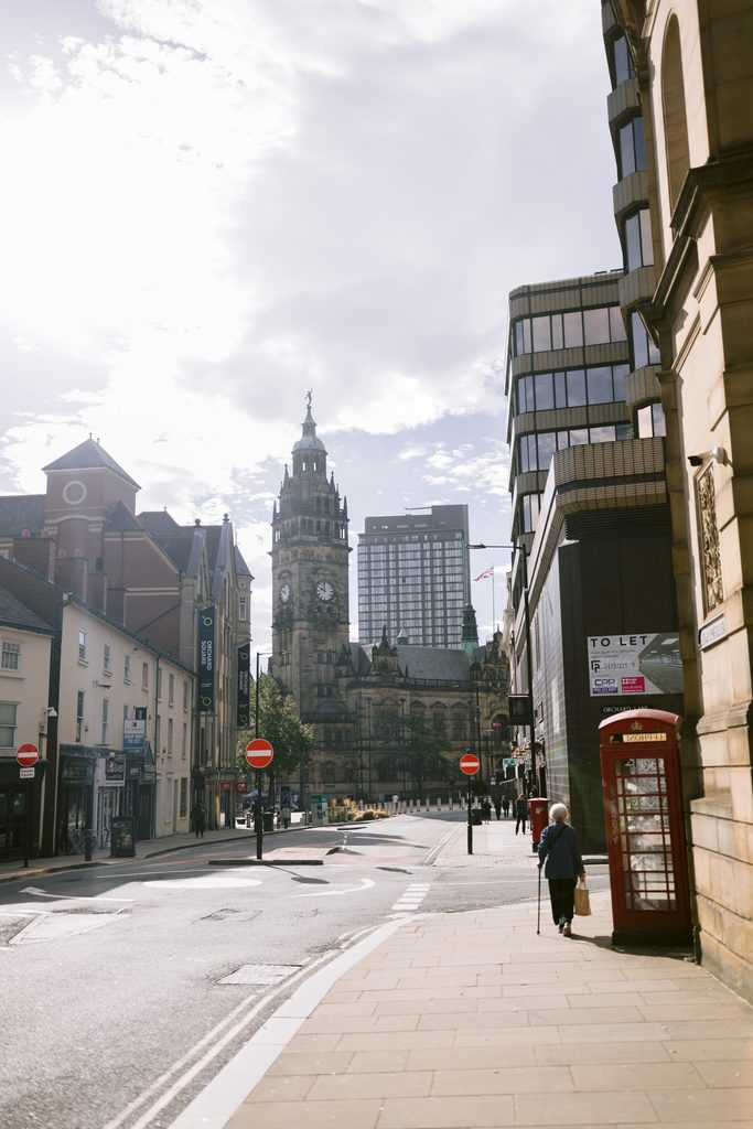 An elderly person walks with a walking stick past a red telephone box on a city street towards a Sheffield Town Hall wedding venue clock tower.