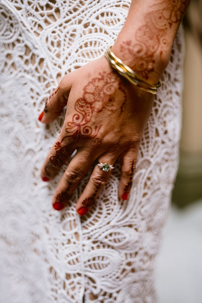 A hand with intricate henna patterns, a gold ring with a green stone, and two gold bangles resting against a white lace dress at a Sheffield Town Hall wedding.