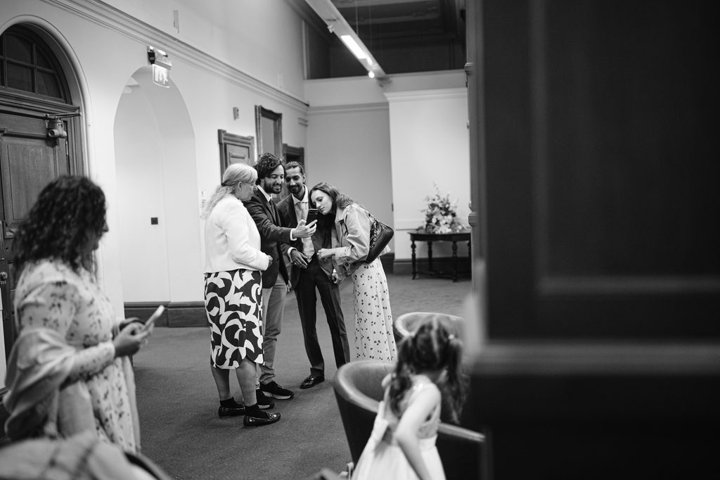 Three people in formal attire look at a mobile phone held by a man in a suit during an intimate wedding at Sheffield Town Hall, whilst a woman and child in the foreground face away from the group.