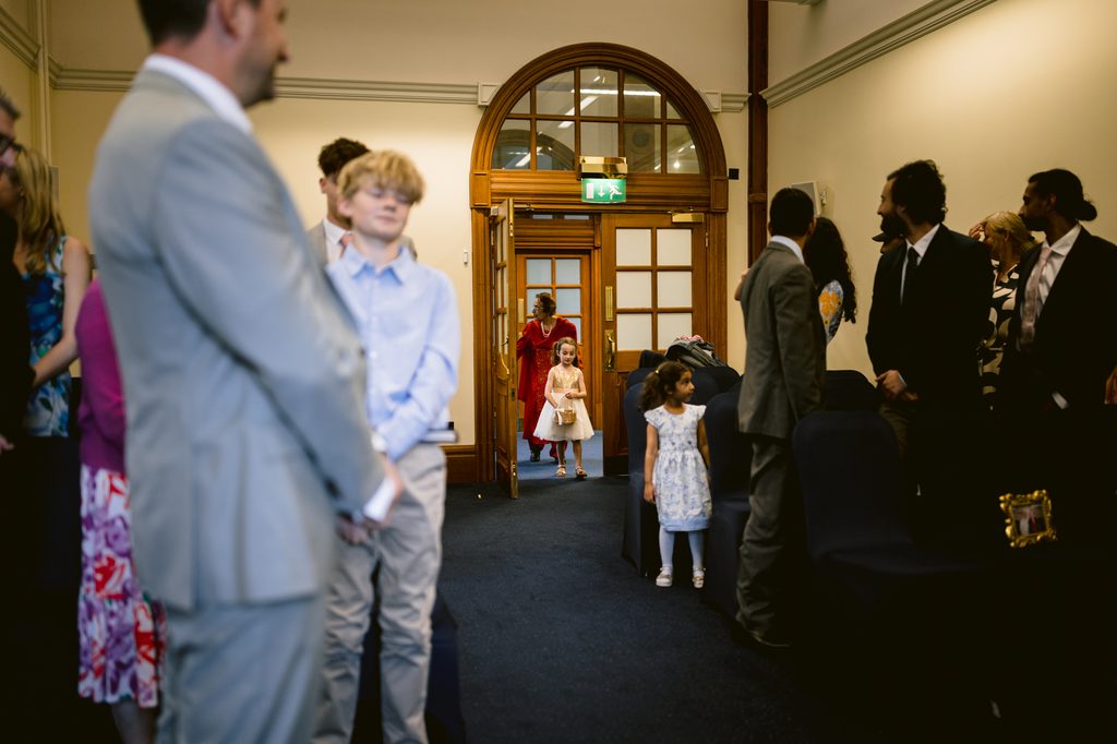 A young flower girl in a white dress walks through a doorway into an intimate Sheffield Town Hall wedding ceremony filled with seated guests.