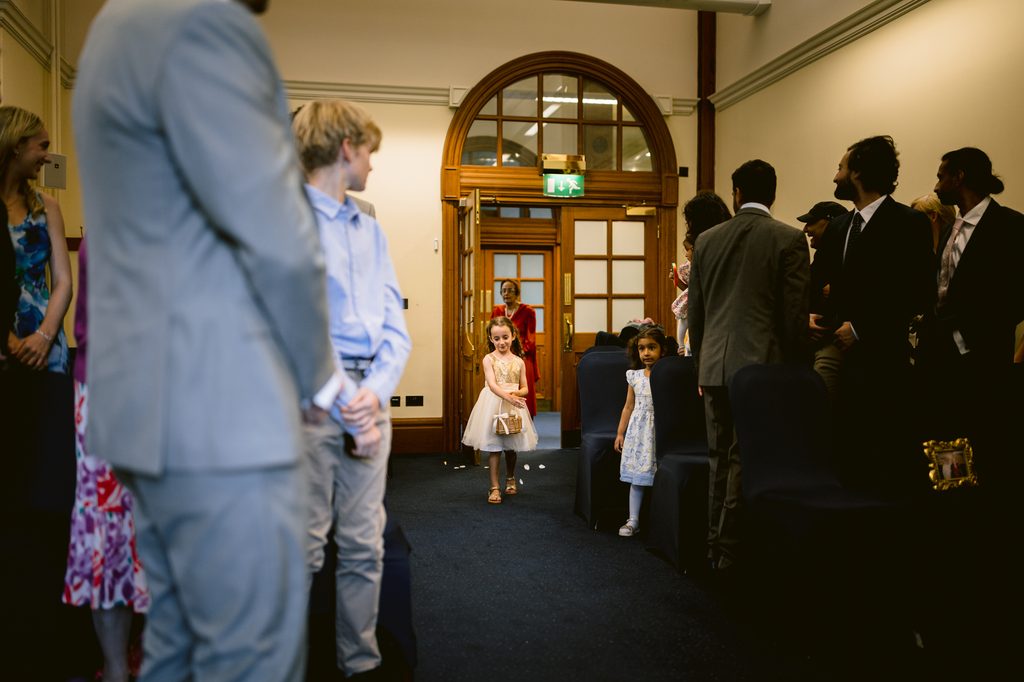 A young flower girl walks down the aisle between rows of wedding guests towards the camera during an intimate wedding at Sheffield Town Hall.