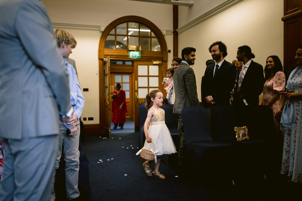 A young flower girl in a white dress walks down the aisle of an intimate wedding, carrying a small basket.