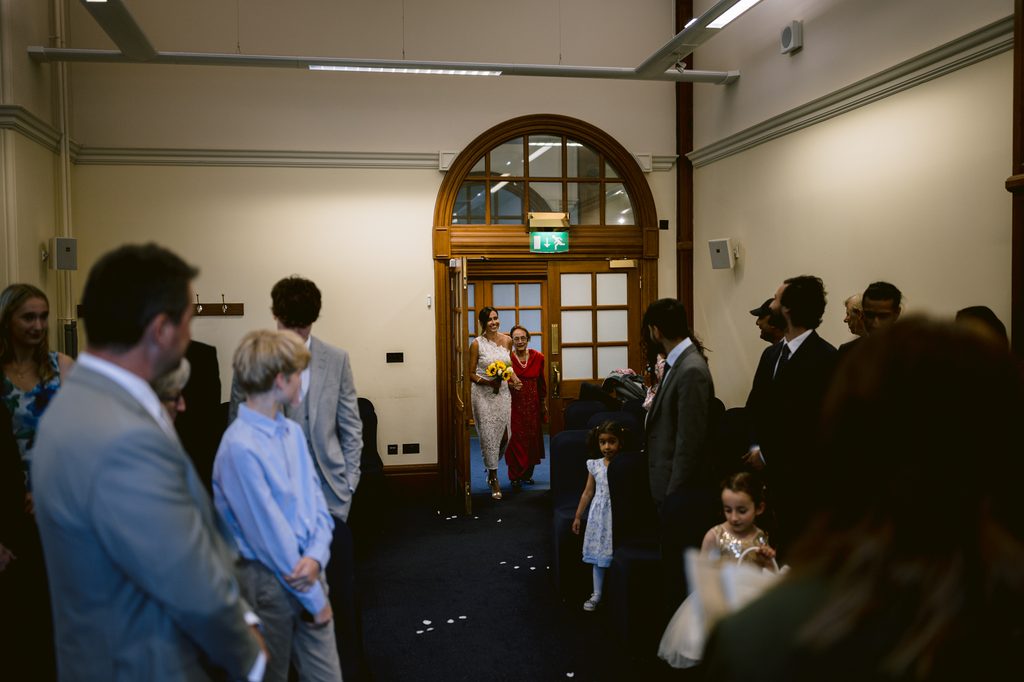 A bride in a white dress holding a sunflower bouquet walks through a doorway at a Sheffield Town Hall wedding, escorted by an older woman in a red dress towards guests seated in the room.