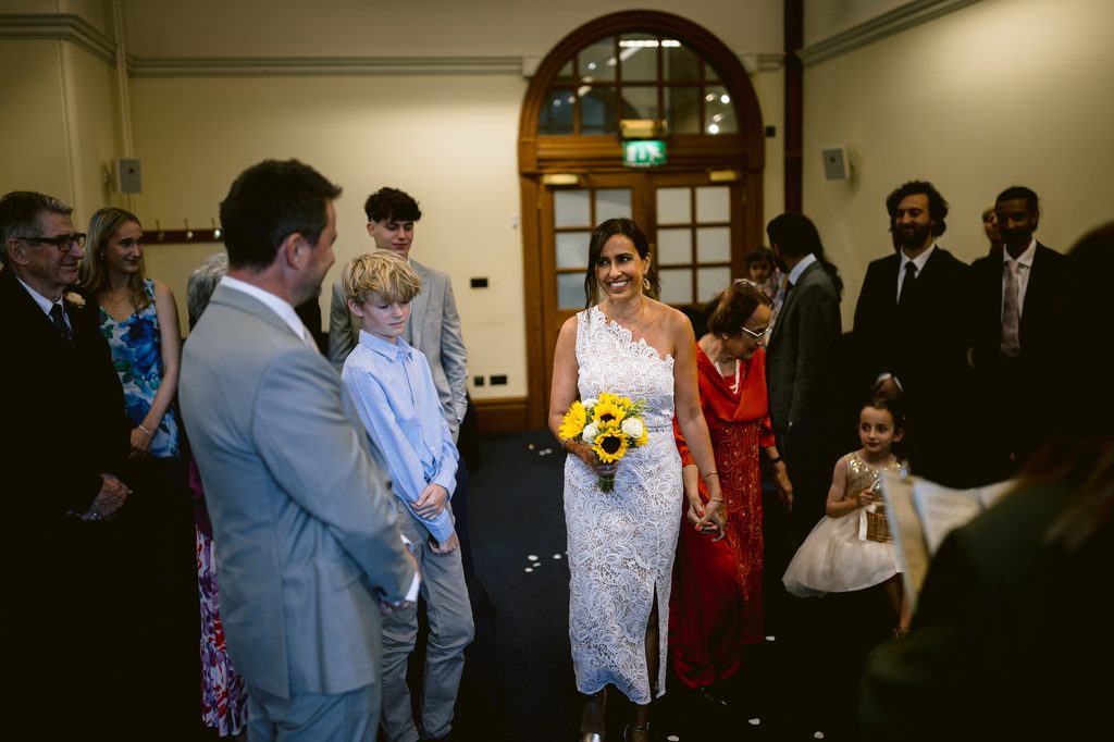 A bride in a white lace one-shoulder dress holding a sunflower bouquet walks down the aisle past guests at an intimate Sheffield Town Hall wedding.