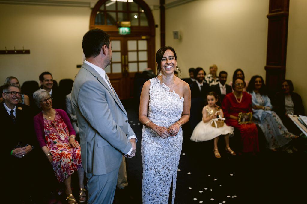 A bride in a white lace dress smiles at her groom in a grey suit during an intimate wedding ceremony at Sheffield Town Hall as guests look on.