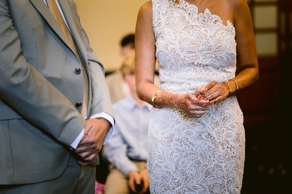 A person in a grey suit stands next to a person in a white lace one-shoulder dress with intricate henna designs on their hands at a Sheffield Town Hall wedding.