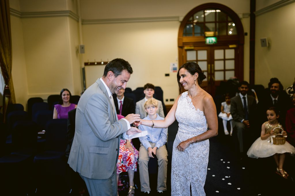 A man in a light grey suit places a ring on the finger of a woman in a white lace dress during an intimate Sheffield wedding ceremony.