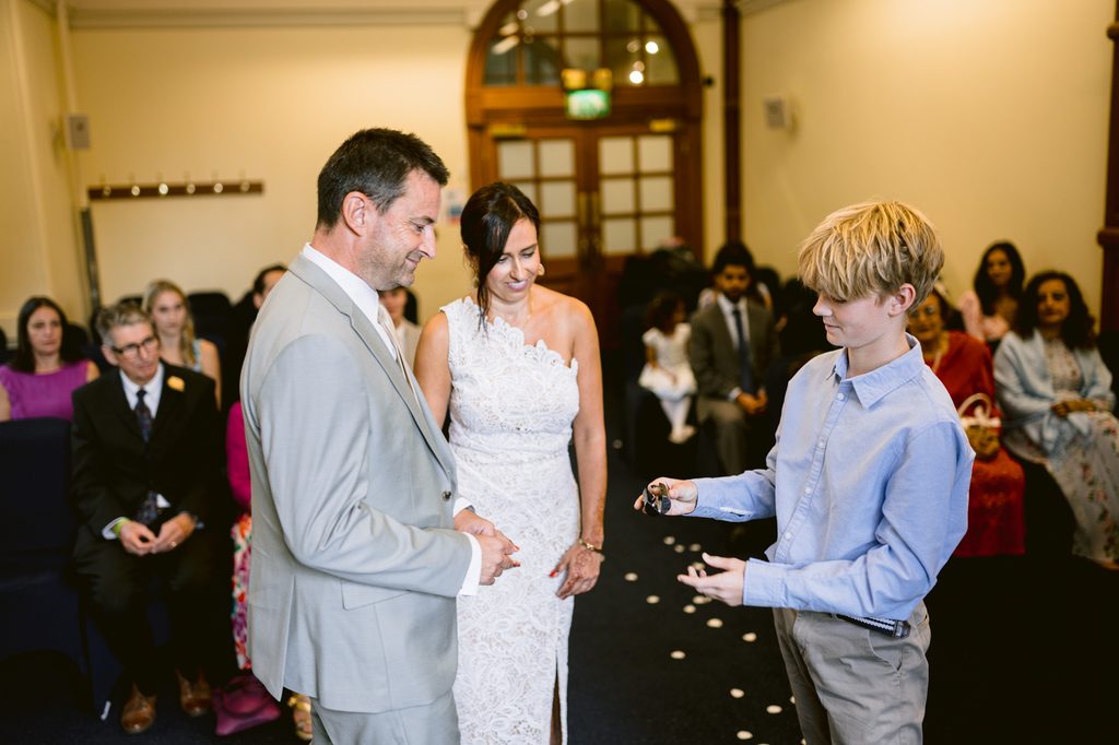 At Sheffield Town Hall, a groom in a light grey suit and a bride in a white lace dress look at a boy in a light blue button-down shirt who is holding wedding rings for this intimate wedding.