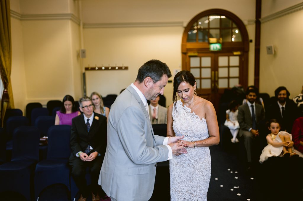 A man in a light grey suit places a ring on the finger of a woman in a white lace wedding dress during an intimate Sheffield Town Hall wedding ceremony.
