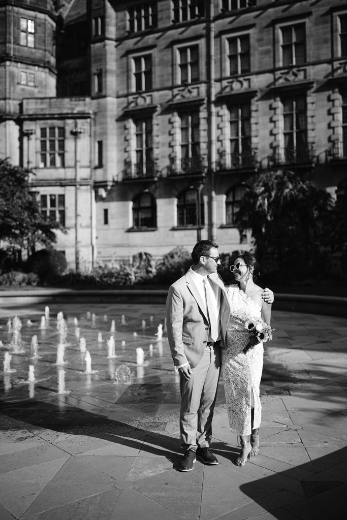 A bride and groom in wedding attire stand together in front of a stone building and a water fountain for an intimate Sheffield wedding.
