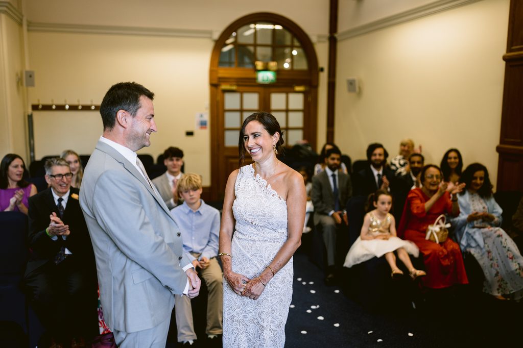 A smiling bride in a white lace dress and a groom in a grey suit look at each other during an intimate Sheffield Town Hall wedding ceremony as guests applaud.