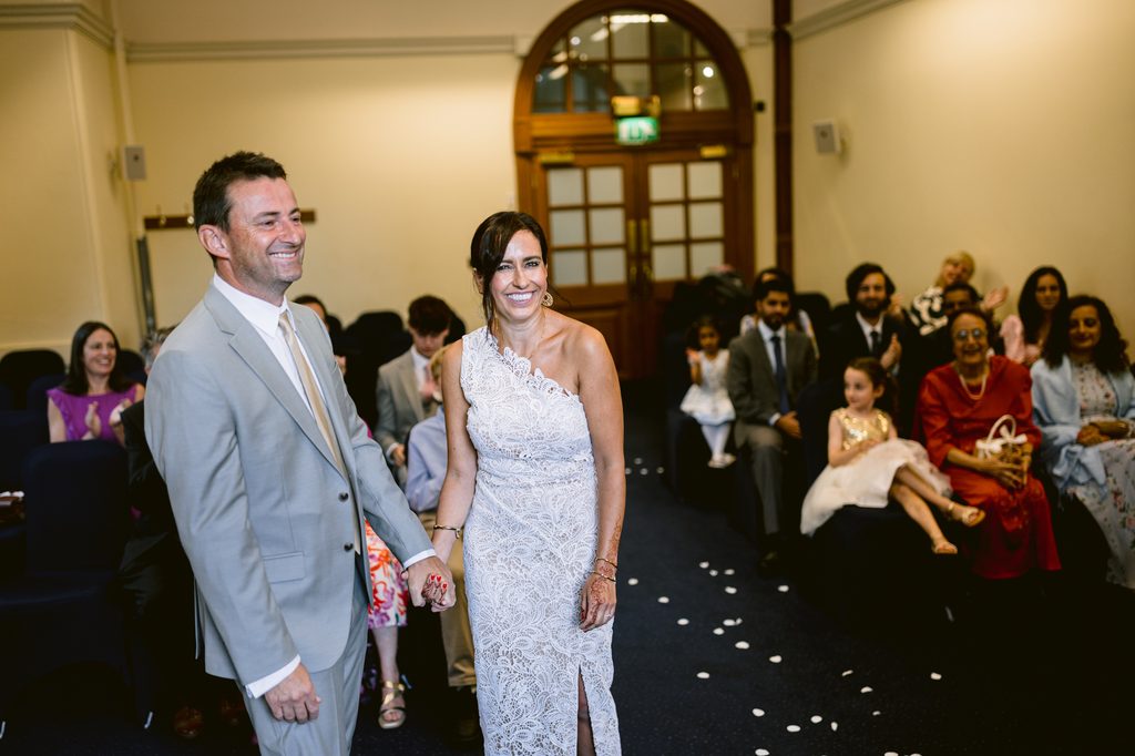A bride in a white lace dress and a groom in a grey suit holding hands, walking down the aisle after their intimate Sheffield Town Hall wedding.