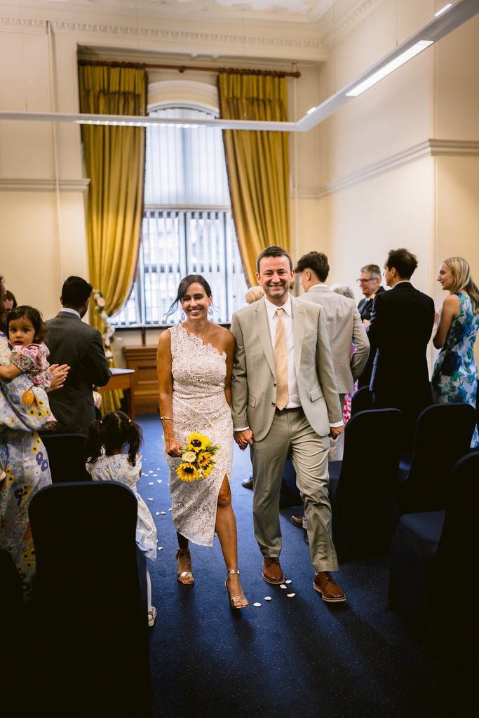 A bride in a white lace dress holding a sunflower bouquet walks down the aisle at a Sheffield Town Hall wedding, hand-in-hand with a groom in a light suit.