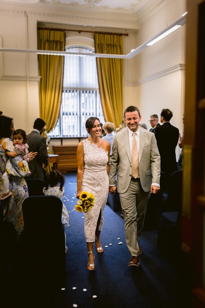 A bride in a white lace dress and a groom in a tan suit walk hand-in-hand down the aisle after their intimate Sheffield Town Hall wedding.